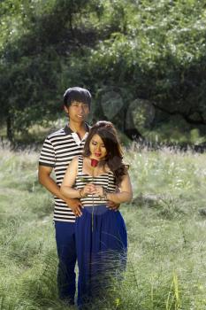 Vertical photo of young adult couple with woman looking at single red rose in the middle of a bright green grass field  