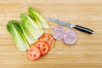 Horizontal photo of freshly cut lettuce, onion and tomato slices on natural bamboo board with kitchen knife