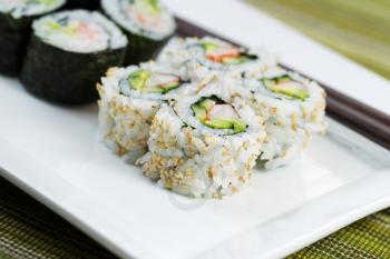 Closeup horizontal photo of freshly handmade California sushi rolls, filling white plate, and chop sticks in background with texture green cloth underneath 
