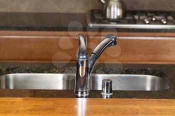 Horizontal photo of a chrome kitchen faucet with stone counter tops, cherry siding and range in background  