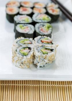 Closeup vertical photo of freshly made California and spicy tuna sushi rolls in white plate and chop sticks in background with bamboo mat underneath 