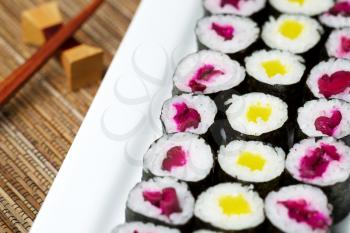 Horizontal photo, focus in middle of plate, of pickled hand rolled sushi on white plate with chop sticks and holder in background and natural bamboo dinner mat underneath