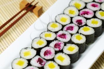 Horizontal photo, focus in middle part of plate, of pickled hand rolled sushi on large white plate with chop sticks and holder in background and natural unstained bamboo dinner mat underneath