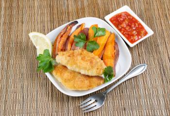 Horizontal top view photo of fried golden breaded coated fish, yams and salsa sauce in white plates with bamboo place mat underneath 