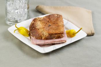 Horizontal photo of ham sandwich made with whole grain bread on white plate with yellow peppers and glass of water in background 