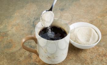 Horizontal photo of a spoon of sugar being poured into a cup of black coffee with stone counter top underneath