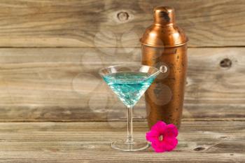 Horizontal photo of a mixed drink, bright pink flower, stir stick and a metal mixer resting on rustic wood