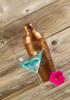 Angled vertical view of a mixed drink, bright pink flower, stir stick and a metal mixer resting on rustic wood