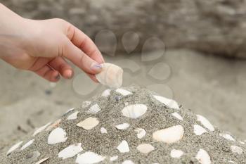 Horizontal photo of female hand placing a seashell on a mound of sand with other shells