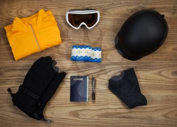 Overhead view of basic ski and snow board accessories placed on rustic wooden boards. Items include helmet, goggles, gloves, sweat shirt, head band, wool socks, and passport.  