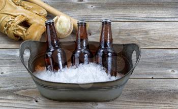  Front view of three brown bottled beers, crushed ice in metal bucket, and baseball equipment in background on rustic wood