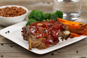 Close up of barbecued spare ribs with yam fries, broccoli, baked beans and glass of water on rustic wooden table. 