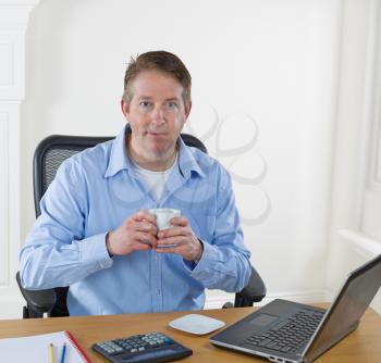Mature man holding cup of coffee, while looking forward, with laptop, calculator, pencils, and notepad on desk. Background is white walls.  