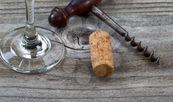 Close up front view of used cork, focus on front part of cork, with antique corkscrew and partial wine glass in background on rustic wood. Horizontal layout. 