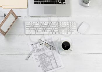 Top view angle of office desktop consisting of laptop, keyboard, pens, mouse, picture frame, phone, coffee, reading glasses, tax forms and work folders on white desk.