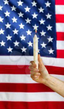 Vertical close up image of a female hand holding a single white candle and glowing flame with United States of America flag in background. Fourth of July holiday concept. 