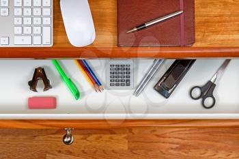 High angle shot of open desk drawer with basic work items inside. Cherry wood desktop has computer keyboard, mouse and executive notepad with pen. Wooden oak floors underneath desk. 