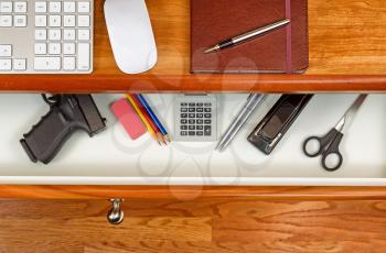 High angle shot of open desk drawer with concealed personal weapon inside. Cherry wood desktop has computer keyboard, mouse and executive notepad with pen. 