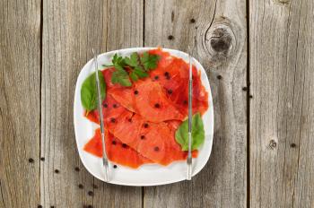 High angled shot of thinly sliced cold smoked red salmon with herbs and seasoning in white plate. Rustic wood underneath. 
