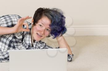 Smiling teen girl, looking forward, giving okay sign while using computer and listening to music at home. 