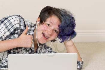 Smiling teen girl, looking forward, giving thumbs up while using computer and listening to music at home. 