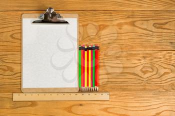 Top view of traditional clipboard, blank paper, ruler, and colorful pencils on wooden desktop. 