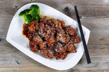 High angled view of Sesame seed chicken with broccoli. Chopsticks on plate with rustic wood underneath.

