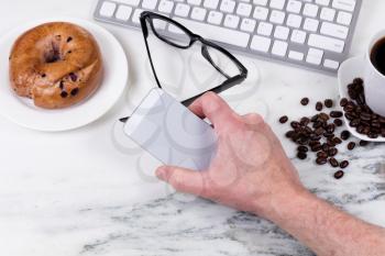 Overhead view marble counter top with male hand holding cell phone with computer keyboard, coffee, reading glasses, and bagel on plate. Work at home concept. Selective focus on upper hand with phone. 