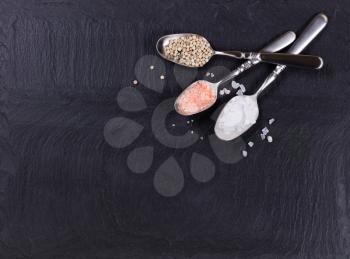 Overhead view of three old spoons containing various spices in upper left hand corner of natural black slate stone. 