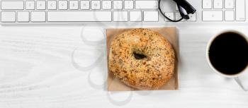Overhead view of a white desktop with keyboard, coffee and bagel. 