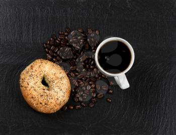 Overhead view of fresh dark coffee, large seeded bagel and roasted beans on natural slate stone. 
