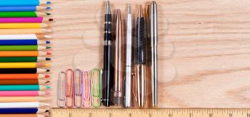 Overhead view of desktop pencils, pens and paper clips with ruler on red oak.  