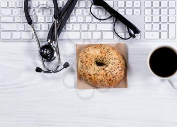 Overhead view of a white desktop with keyboard, stethoscope, reading glasses, coffee and bagel. 