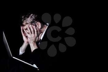 Side view of stressed business man, holding his head, with laptop in front of him.  Dark background with light on subject.  