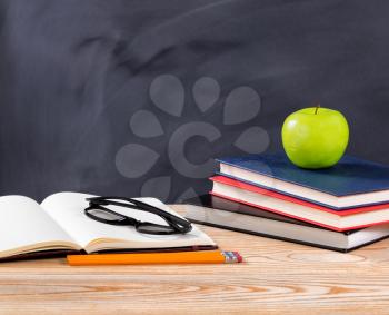 Back to school concept with reading glasses, pencils, notepad, books and green apple on desktop with erased black chalkboard in background. 