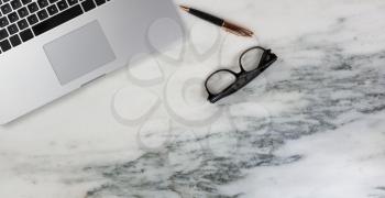 Flat lay of a partial laptop, pen and reading glasses on natural marble desktop
