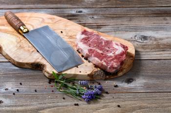 Large butcher knife and raw beef with marbled fat on carving board with spices and herbs