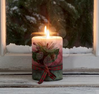 Christmas candle glowing on window sill with snowy evergreen branches in background 