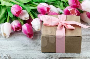 Close up view of a brown gift box with pink tulips in background on white weathered wooden boards 