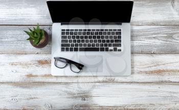 Office desktop setting with laptop, baby plant and reading glasses on white rustic desk