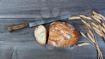Overhead view of baked whole loaf of bread with cutting knife and wheat stalks on rustic wooden boards