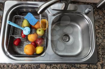 Fresh vegetables and fruits being washed in kitchen sink. Primary focus on food with running water from faucet.  