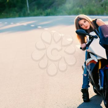 Young beautiful blond woman in fashionable jeans and a black T-shirt looks into the mirror behind the wheel of a modern motorcycle. Outdoor portrait in soft sunlight.