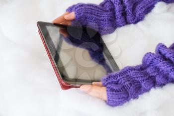 Woman hands in light teal knitted mittens are holding modern tablet pc on snow background. Winter concept.