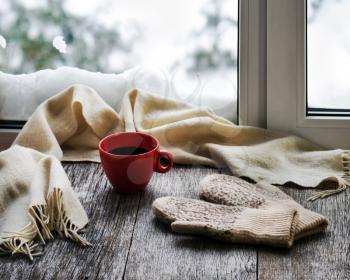 Red cup of coffee or tea, beige scarf and womans mittens are located on stylized wooden windowsill. Winter concept of comfort and relaxation.