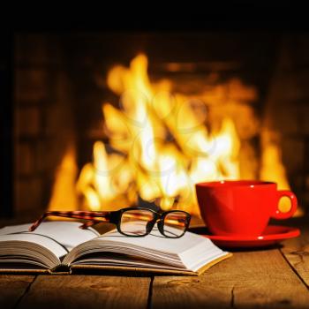 Red cup of coffee or tea, glasses and old book on wooden table near fireplace. Winter and Christmas holiday concept.