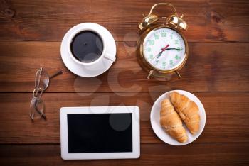 Coffee, alarm clock, glasses, digital tablet and croissants on the wooden table, top view.