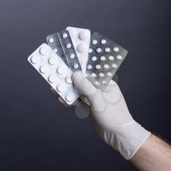 Male hand in glove holding pills in blisters on dark background. Square format.
