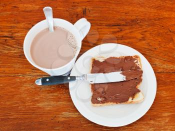 sweet sandwich from fresh toast with chocolate spread, table knife and cup of hot chocolate on wooden table