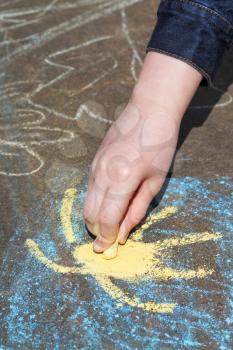 girl paints yellow sun in blue sky with colored chalk on asphalt outdoors